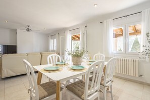 A dining area with a white table set for six, surrounded by white chairs with brown cushions. A ceiling fan is visible above, and a radiator is on the wall to the right.