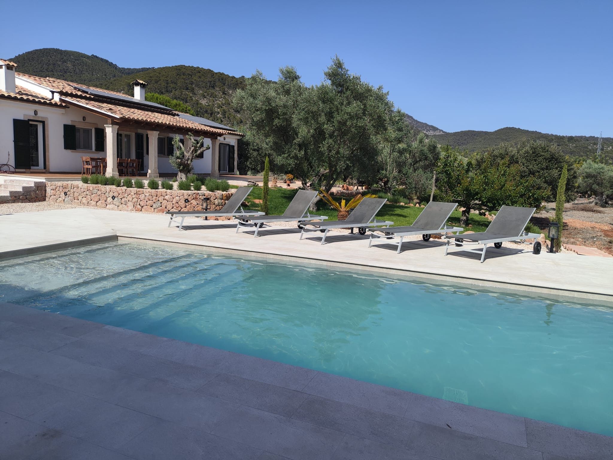Una piscina con sillas de descanso, un muro de piedra y una casa con tejado de baldosas.