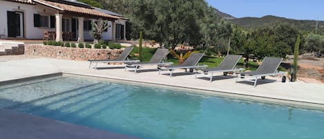 Una piscina con sillas de descanso, un muro de piedra y una casa con tejado de baldosas.