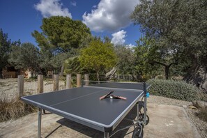 Una mesa de ping pong al aire libre con una pelota y paletas, rodeada de árboles y un cielo despejado.