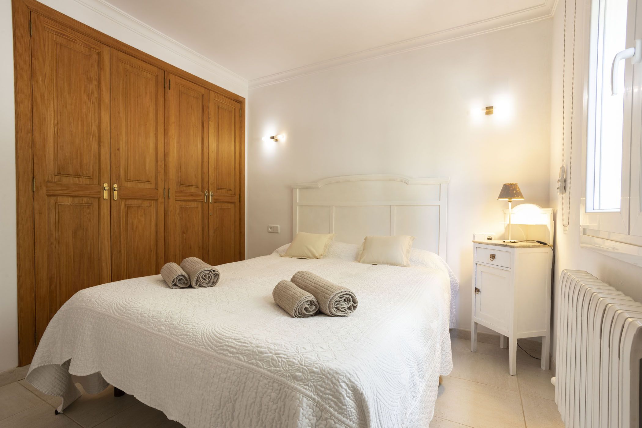 A bedroom with a large wooden wardrobe, a white bed with rolled towels, a bedside table with a lamp, and a radiator.