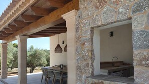An outdoor patio with a stone wall, a wooden pergola, a table with chairs, and hanging lights. - 4 bedroom country house with pool and barbecue (Bunyola)