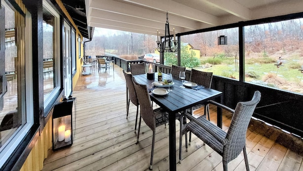 En træterrasse med et spisebord, store vinduer med udsigt over en have, og en hængende lampe.