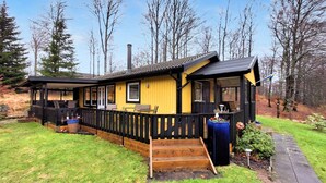 En gul hytte med træterrasse, overdækket veranda og skorsten.