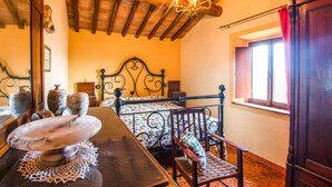 A bedroom with a wooden ceiling, a bed with a floral bedspread, a wooden nightstand with a white ceramic pot, and a window with a red curtain. - Lovely apartment in Volterra with WiFi (Volterra)