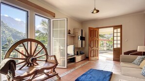 Un vaste salon comprenant un grand fauteuil à bascule en bois, un canapé et un téléviseur posé sur un meuble en bois. Une grande fenêtre offre une vue sur les montagnes et la verdure.