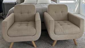 Une chambre à coucher comprenant un lit, deux fauteuils, un ventilateur de plafond et de grandes fenêtres équipées de stores.