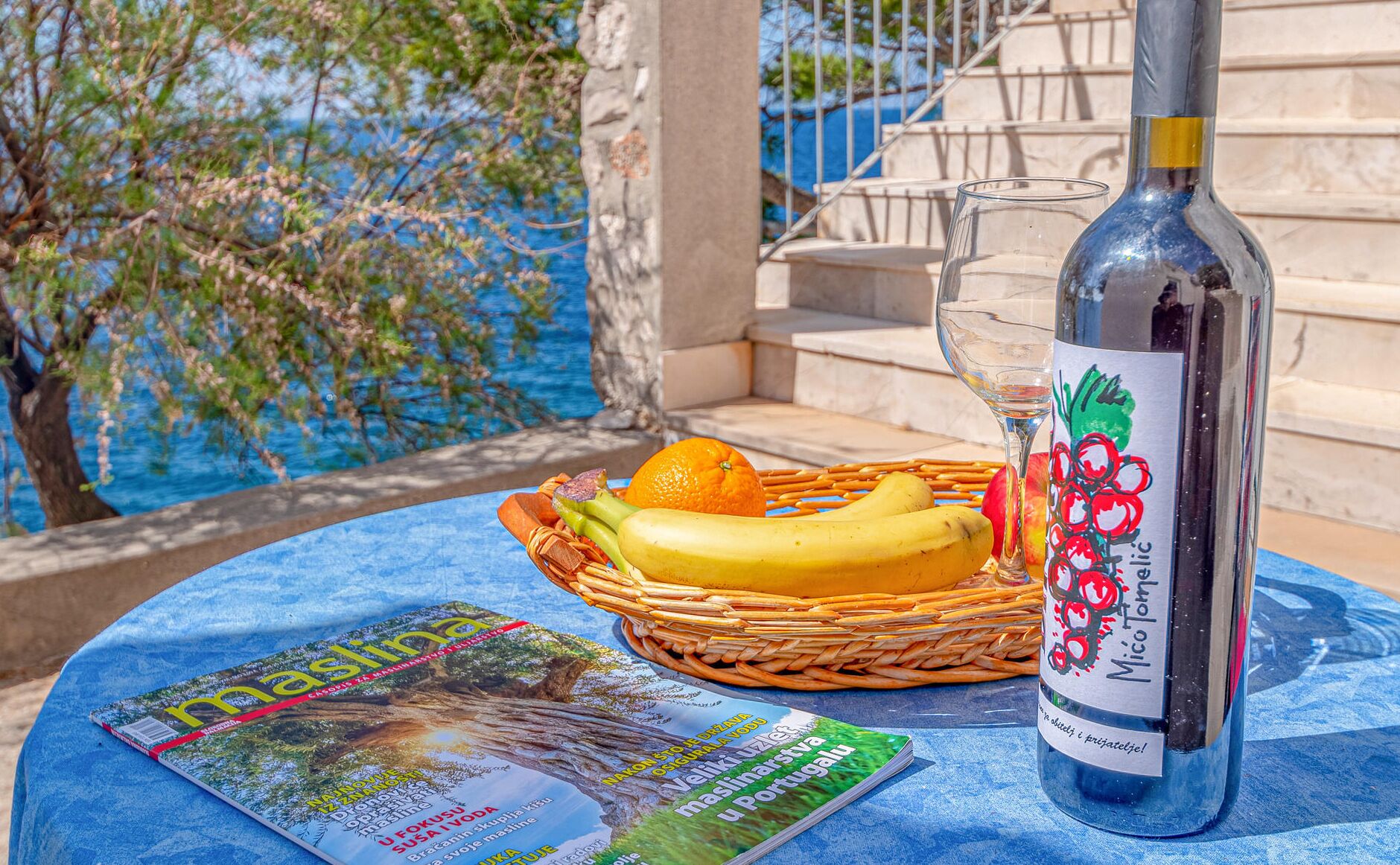 Een tafel met een fles Mico Tomelic wijn, een glas, een mand met fruit en een tijdschrift.