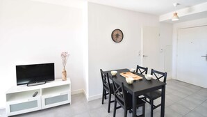 Un salon moderne avec un téléviseur à écran plat posé sur un meuble de rangement blanc, une table à manger avec des chaises et une décoration minimaliste comprenant un tableau mural représentant une boussole.