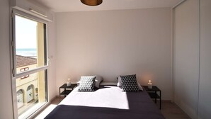 Une chambre avec une grande fenêtre, un lit avec deux oreillers, une table de chevet avec une bougie et un luminaire suspendu.