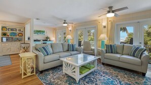 A spacious living area with a beige sofa set, a glass coffee table, and a patterned rug. There are ceiling fans, a kitchen area in the background, and large windows with curtains. - Bay 2 Bay: Bay Across Street! Amazing Views of Bay & Canal, Pool & Lounge by Anna Maria Life (Anna Maria)