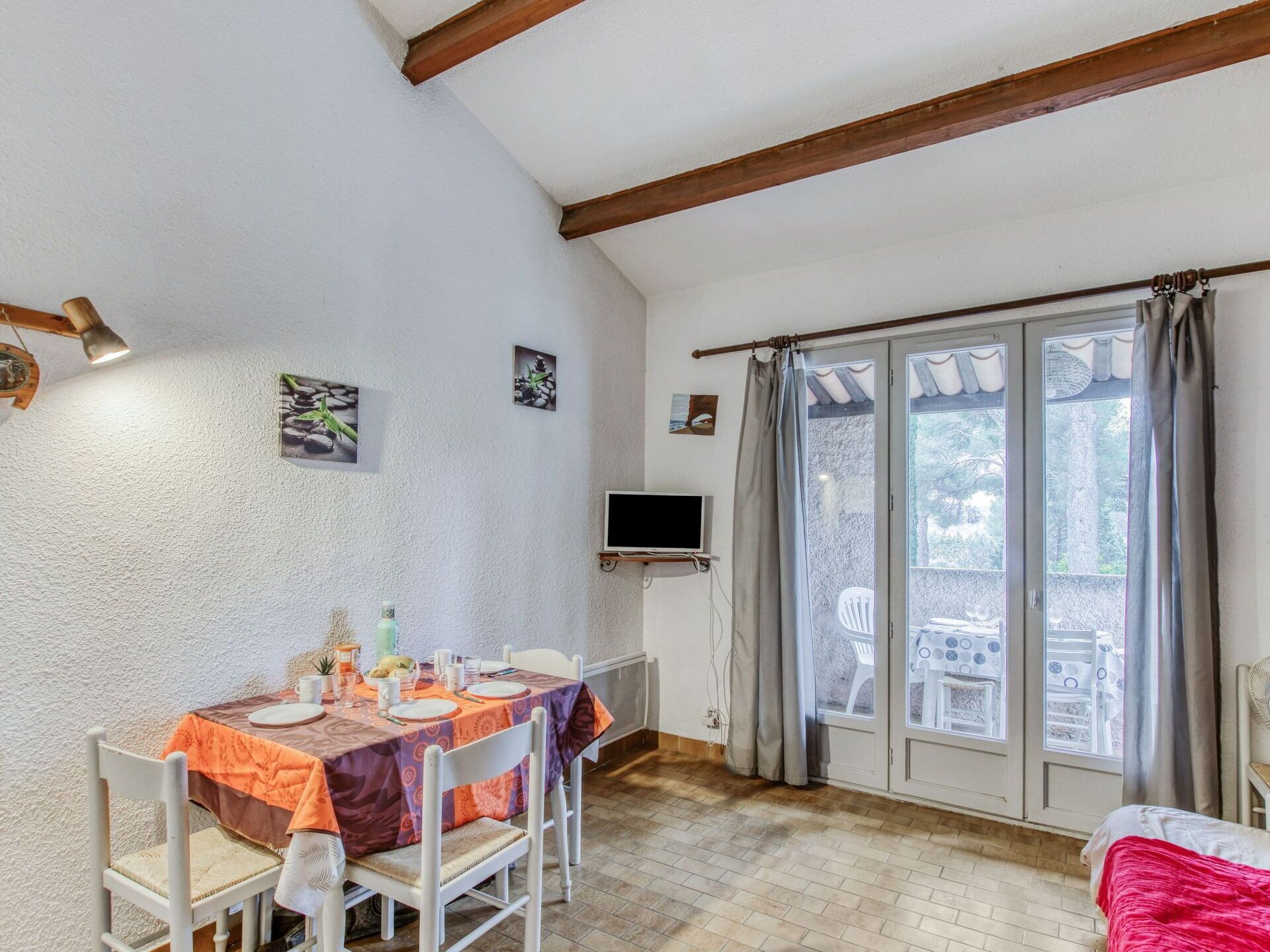 A dining area with a table set for two, a television mounted on the wall, and a window with curtains.