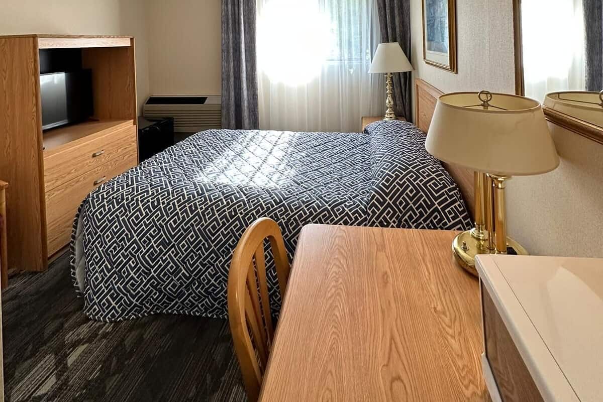 A hotel room with a bed, a desk, a chair, a television, a lamp, and a large mirror.