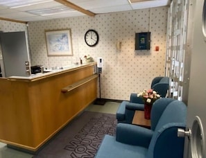 A reception area with a wooden desk, a clock, a wall-mounted control panel, and a seating area with a floral arrangement.