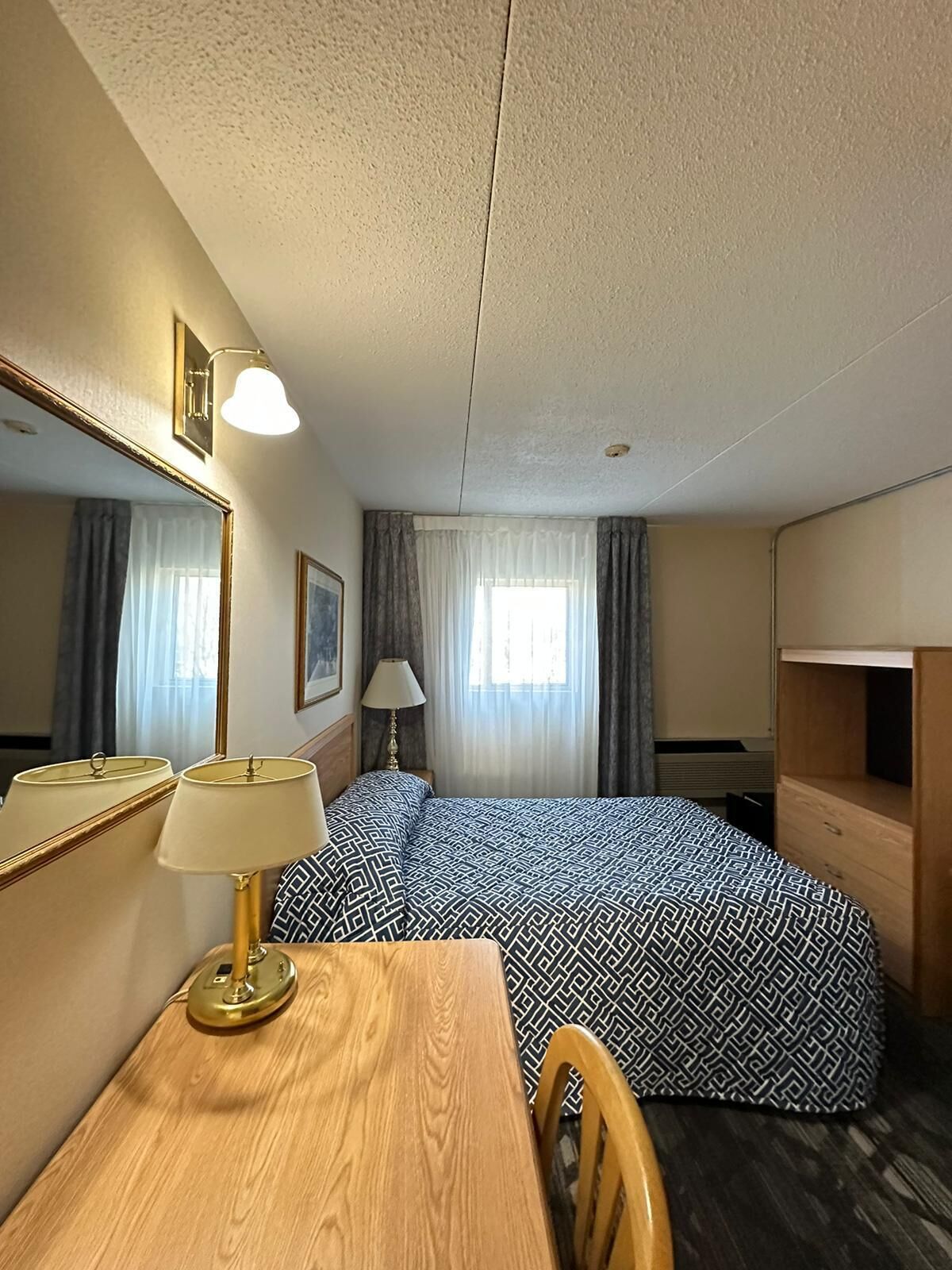 A hotel room with a bed, a desk, a chair, a lamp, a mirror, and a window with curtains.