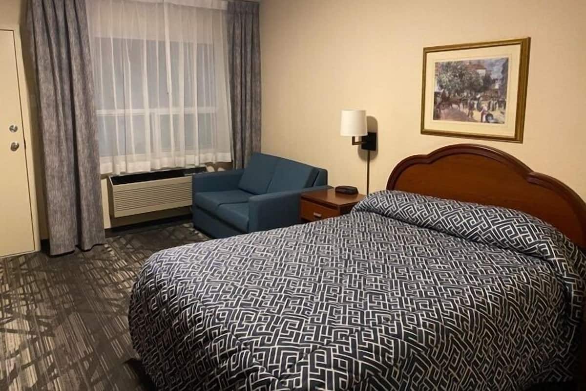 A hotel room with a bed, a chair, a lamp, and a painting on the wall.