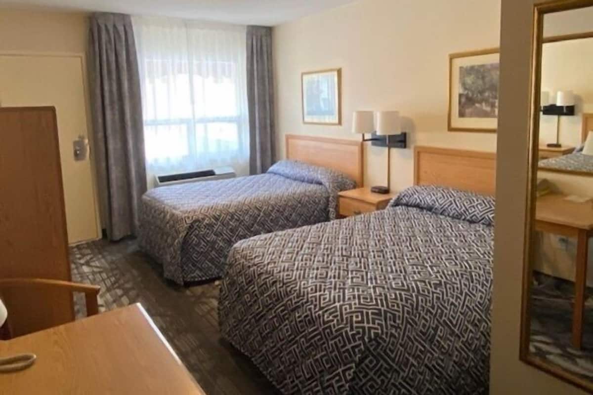A hotel room with two beds, a desk, a chair, a window with curtains, a lamp, and a framed picture on the wall.