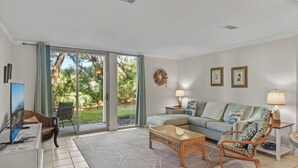 A living room with a sofa, a chair, a coffee table, a TV, and a view of greenery through large sliding doors.