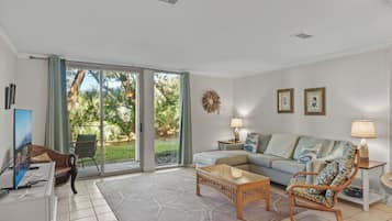 A living room with a sofa, a chair, a coffee table, a TV, and a view of greenery through large sliding doors.