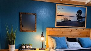 A bedroom with a bed, a nightstand, a lamp, a framed picture, and a shelf with plants and a sign. - Fish Cabin at the Pines - Lake O' the Pines - Hot Tub - Fire Pit (Jefferson)