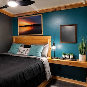 A bedroom with a bed, a nightstand with a lamp, a framed picture, and a potted plant. - Fish Cabin at the Pines (Jefferson)