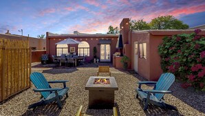 Un patio con sillas Adirondack, un hogar para fogata y una casa de fachada roja y ventanas arqueadas.