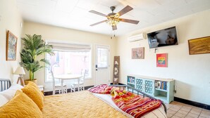 Un dormitorio bien iluminado con cama, ventilador de techo, televisor, estantería y ventana con persianas.