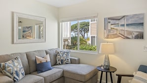 A living room with a grey sectional sofa, a round coffee table, and a lamp. There is a large window with a view of a beach and a house. - LOWER Unit at Niguel Beach Terrace, Dana Point!  Close to Pool  view (Dana Point)
