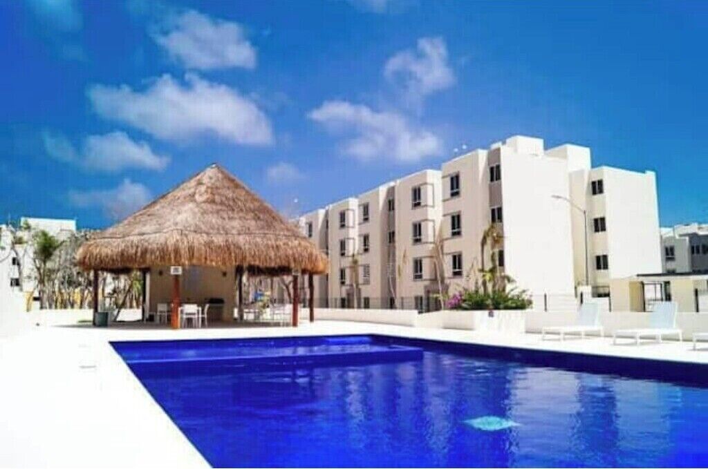 A swimming pool with a thatched-roof structure, modern buildings, and clear skies.
