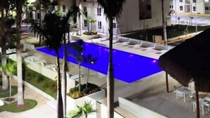 A swimming pool with a blue-lit edge, surrounded by palm trees and a multi-story building with balconies. - Rouse's House near Hotel Zone, Zamna Fest (Tulum)