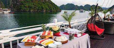 A table set with various fruits and snacks on a boat deck with a view of a mountainous island and the sea.
