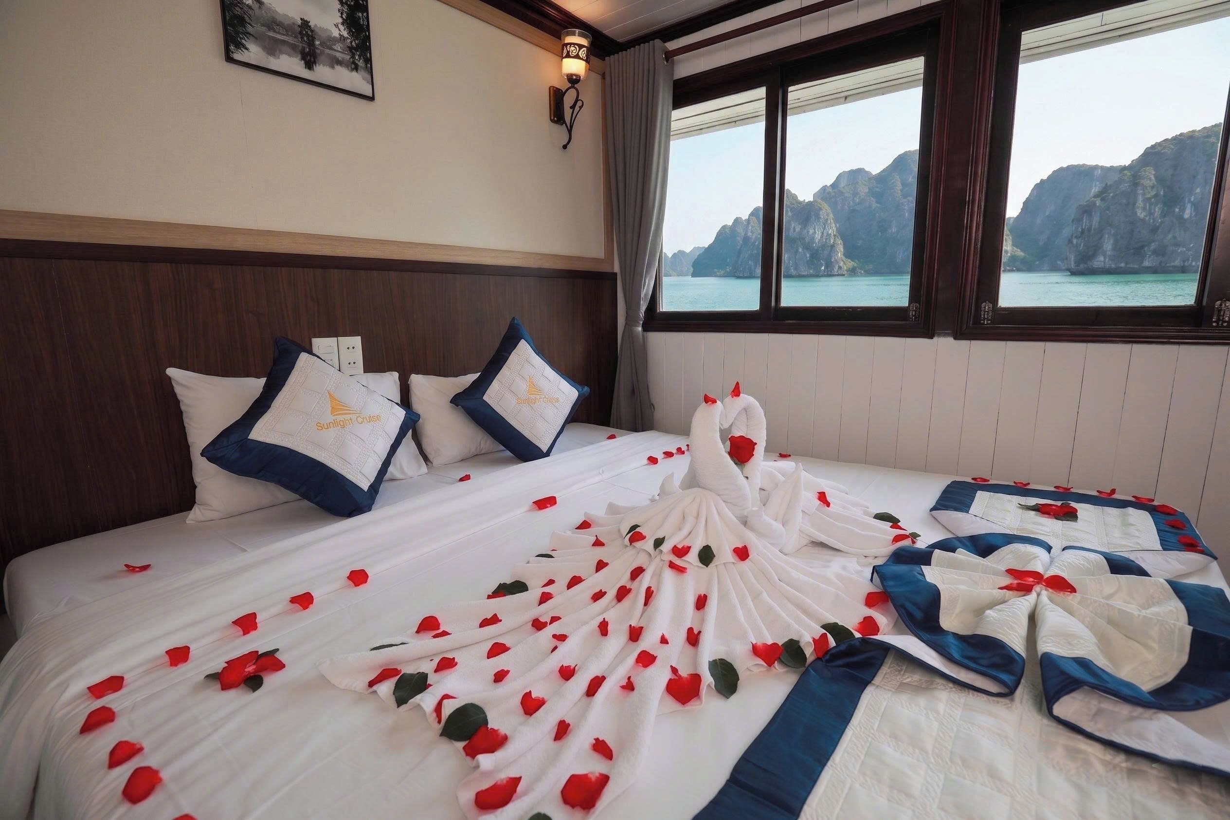 A hotel room with a bed adorned with rose petals and a swan-shaped pillow, a view of a mountainous coastline, and a framed picture on the wall.
