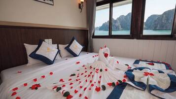 A hotel room with a bed adorned with rose petals and a swan-shaped pillow, a view of a mountainous coastline, and a framed picture on the wall.