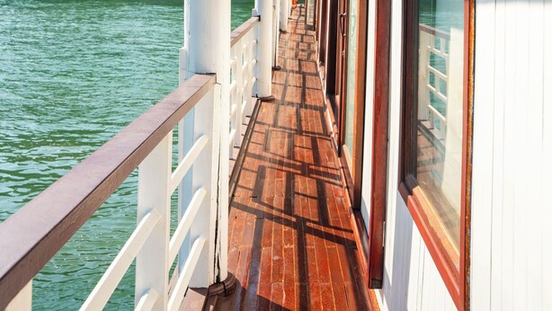 Een houten dek met witte reling op een boot, met uitzicht op kalm water en kliffen.