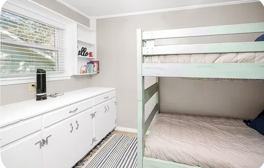 A bunk bed with a white and green frame, a white dresser with drawers, a radiator, and a window with blinds.