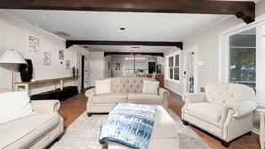 A spacious living room with a large beige sofa, a matching armchair, and a coffee table. There is a wooden ceiling beam, a chandelier, and a view into the kitchen.