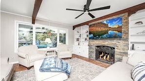 A living room with a fireplace, a ceiling fan, a large TV, and a view of the outdoors.