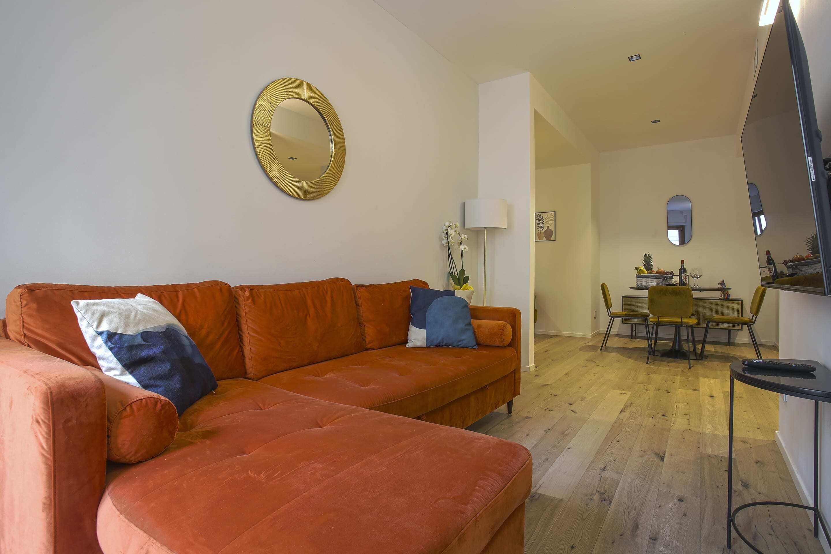 Ein Wohnzimmer mit einem Ecksofa, einem runden Spiegel, einem Essbereich mit Tisch und Stühlen sowie einem Fernseher auf einem Lowboard.