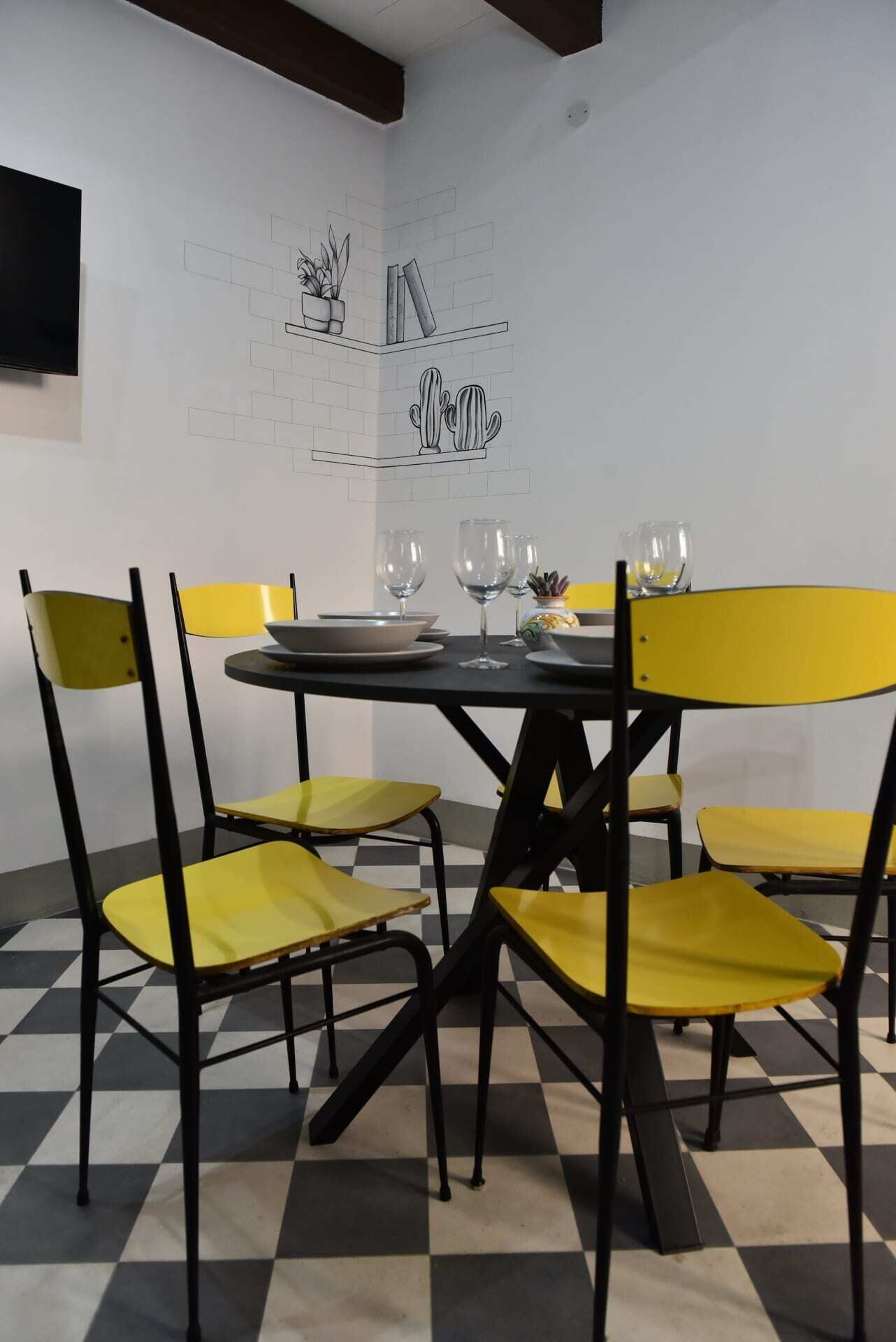 A dining area with a black table and yellow chairs, a black and white checkered floor, and a wall with a cactus and plant illustration.
