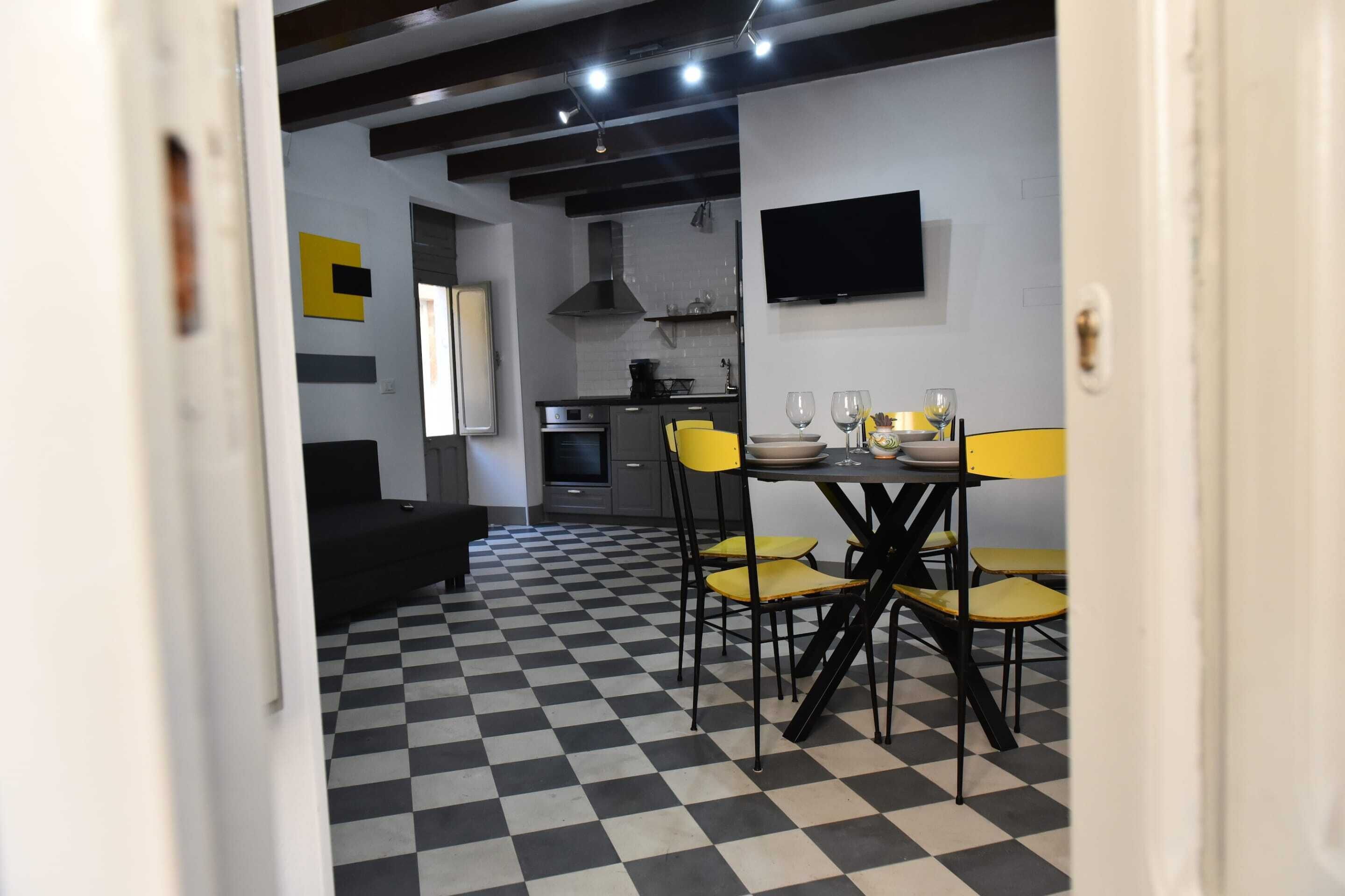 A compact kitchen with modern appliances, a dining area with yellow chairs, and a checkered floor.