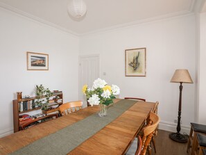 Ein Esszimmer mit Holztisch, Stühlen, einer Blumenvase, einem Bücherregal und zwei gerahmten Bildern an der Wand.