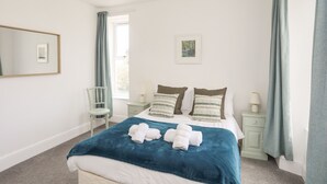 Une chambre à coucher comprenant un lit, deux tables de chevet, une chaise, une fenêtre avec des rideaux, un luminaire suspendu et un tableau encadré au mur.