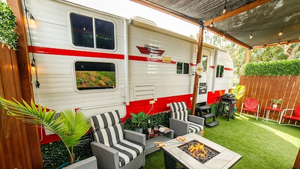 A recreational vehicle with a red stripe, outdoor seating with striped cushions, and a fire pit.