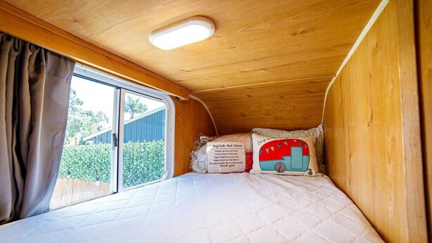 A small, cozy sleeping area with a bed, a pillow with a camper van design, and a sign with care instructions. - End of Summer Savings at Darling Daisy! (El Cajon)