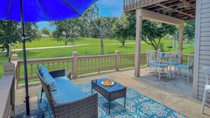 Eine überdachte Terrasse mit Korbmöbeln, einem blauen Sonnenschirm und Blick auf einen Golfplatz.