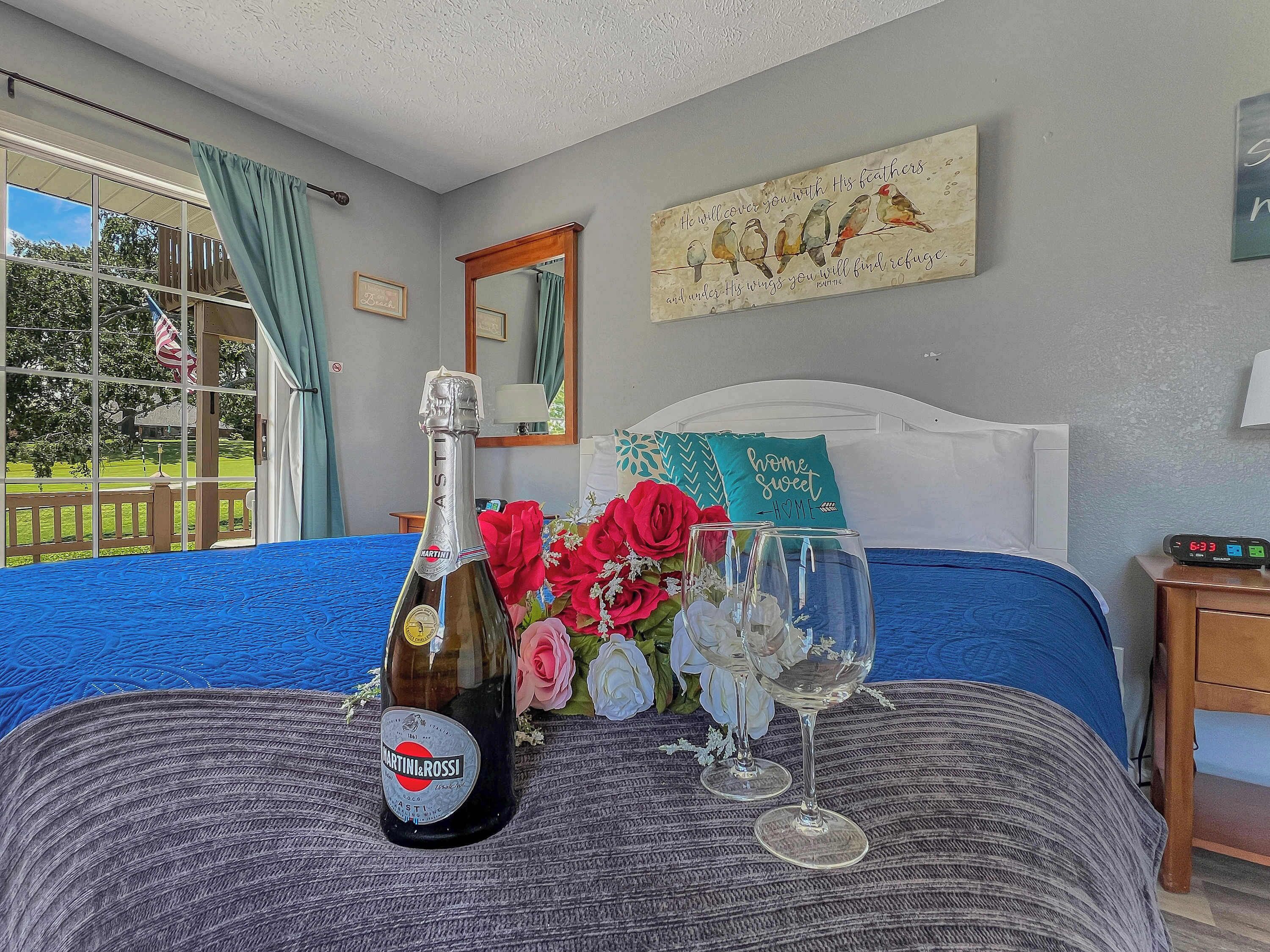A bedroom with a bed, a bottle of Martini & Rossi wine, two wine glasses, and a bouquet of flowers.