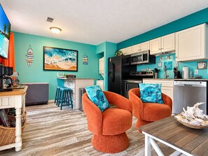 A compact living space with teal walls, a kitchen area, and two orange armchairs with blue cushions.
