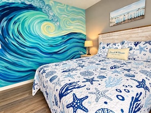 A bedroom with a large wave-themed mural, a bed with starfish and shell patterns, and a painting of birds on a fence.