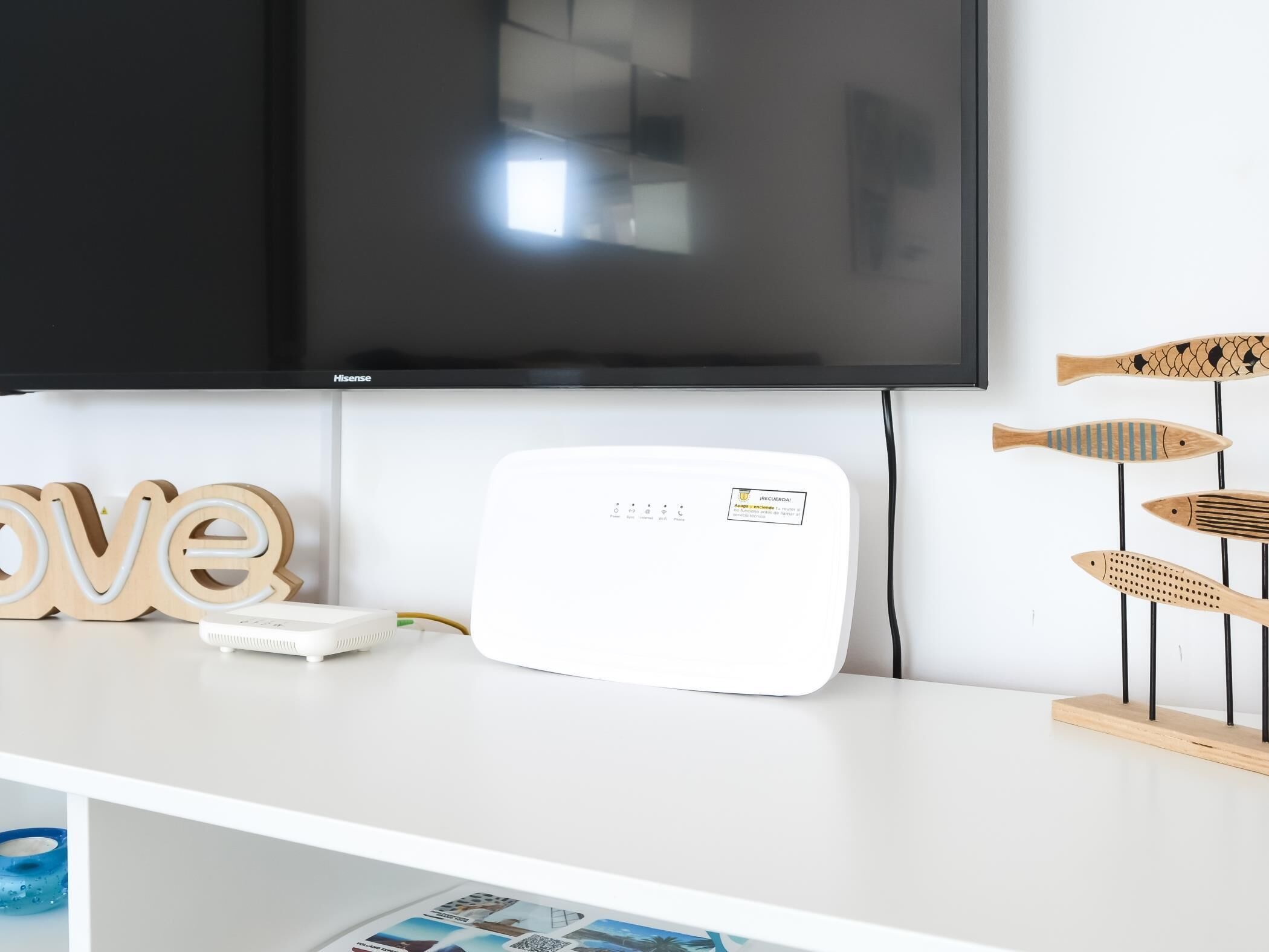 Un téléviseur à écran plat fixé au mur, un appareil électronique blanc posé sur une surface blanche, des décorations en bois en forme de poisson et un panneau en bois sur lequel est écrit le mot « amour ».
