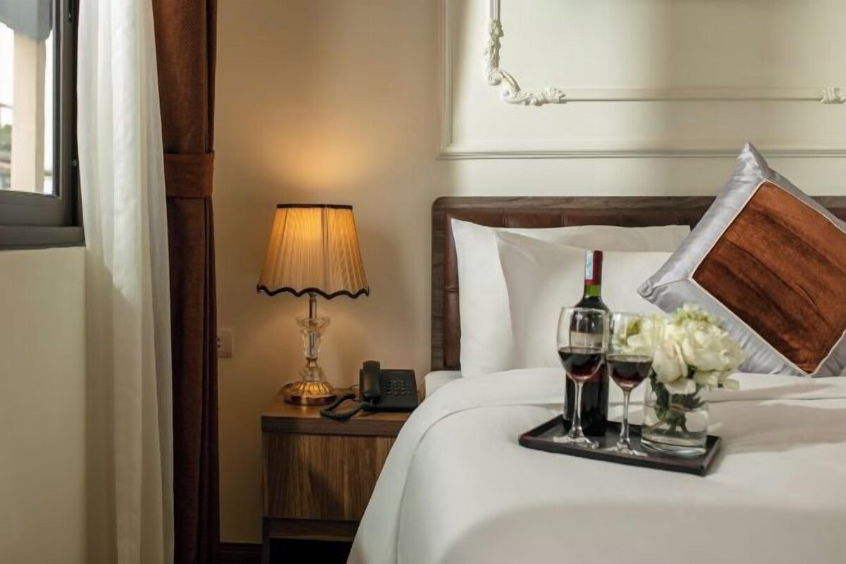 A neatly made bed with a tray of wine glasses and flowers, a lamp, and a telephone on a wooden nightstand.